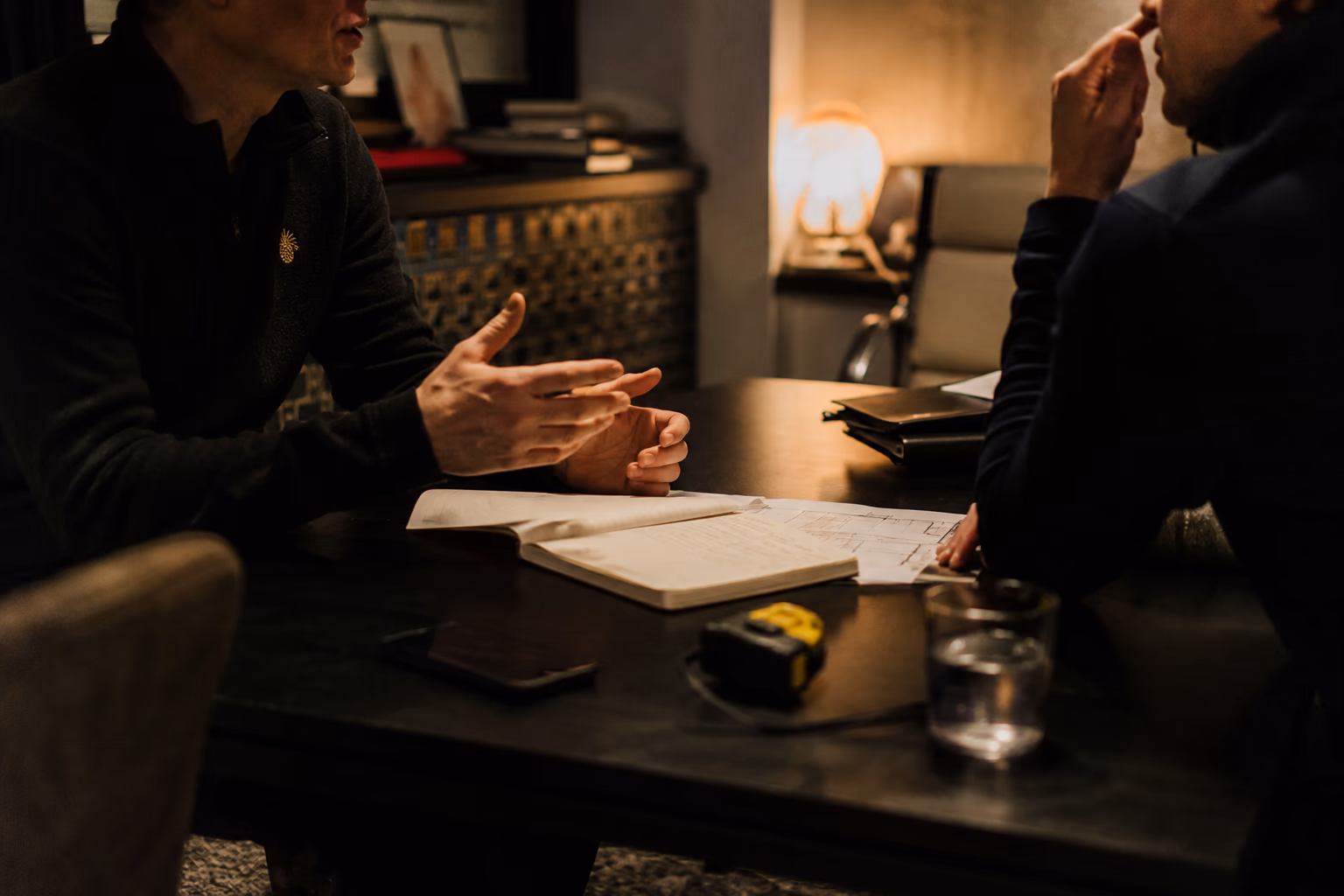 Deux personnes en discussion autour d'une table, un carnet ouvert, le plan du lieu et un mètre ruban posés dessus
